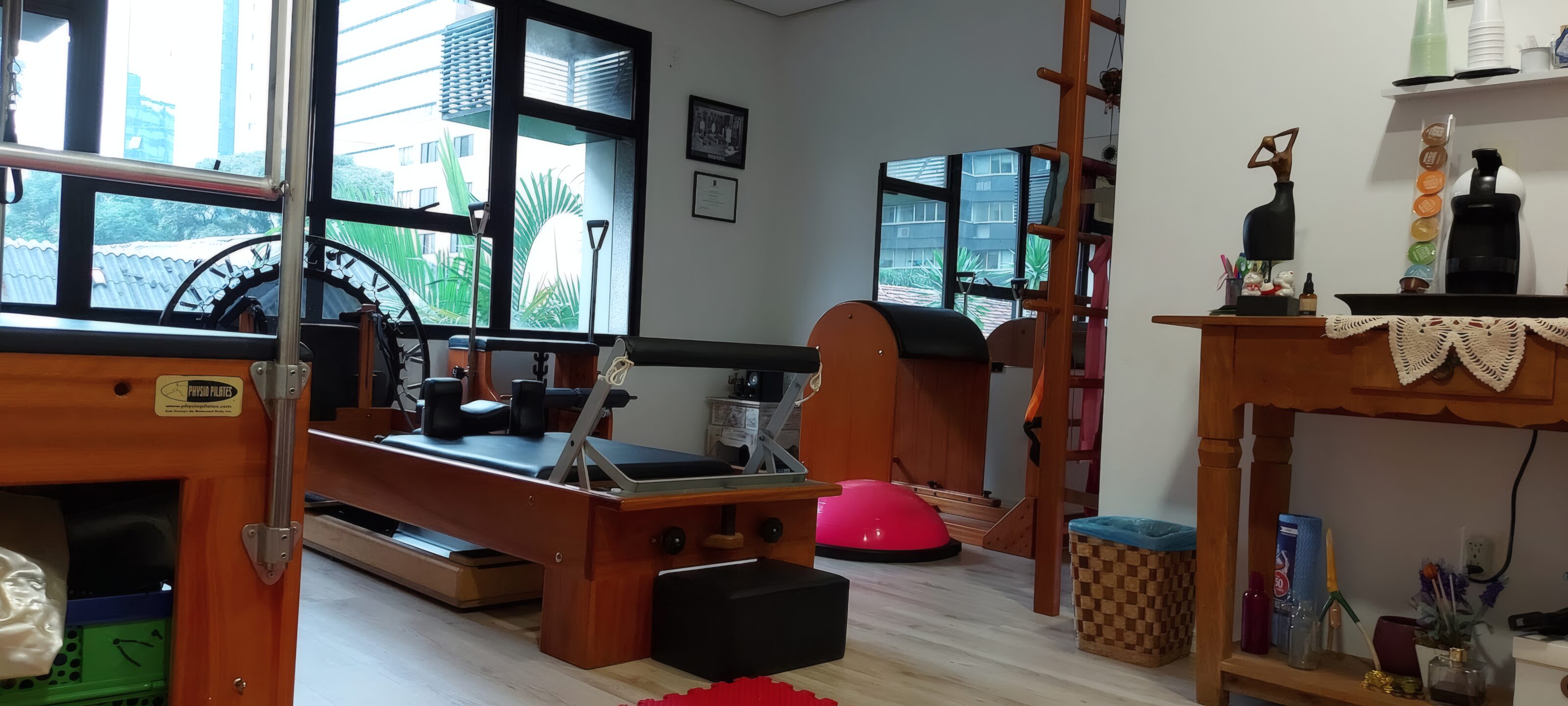 Sala do estúdio com reformer, barrel e escada de madeira, e a vegetação do bairro pela janela